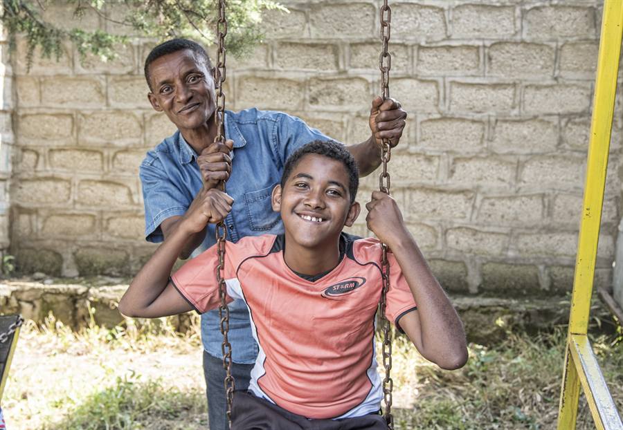 A father smiles as he pushes his beaming son on a swing in a sunny playground.