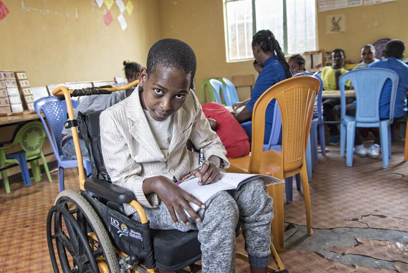 A boy seated in his wheelchair writes carefully in his exercise book during class, while other learners work at tables behind him.