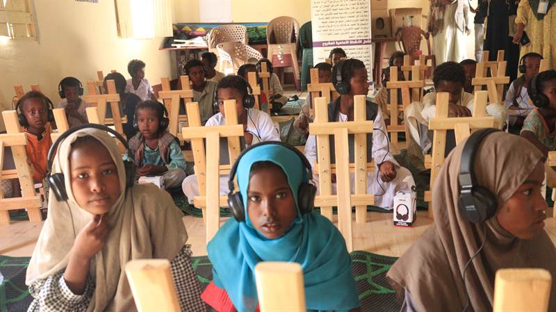 Children with headphones learn on tablets mounted on wooden easels at an e-learning centre.