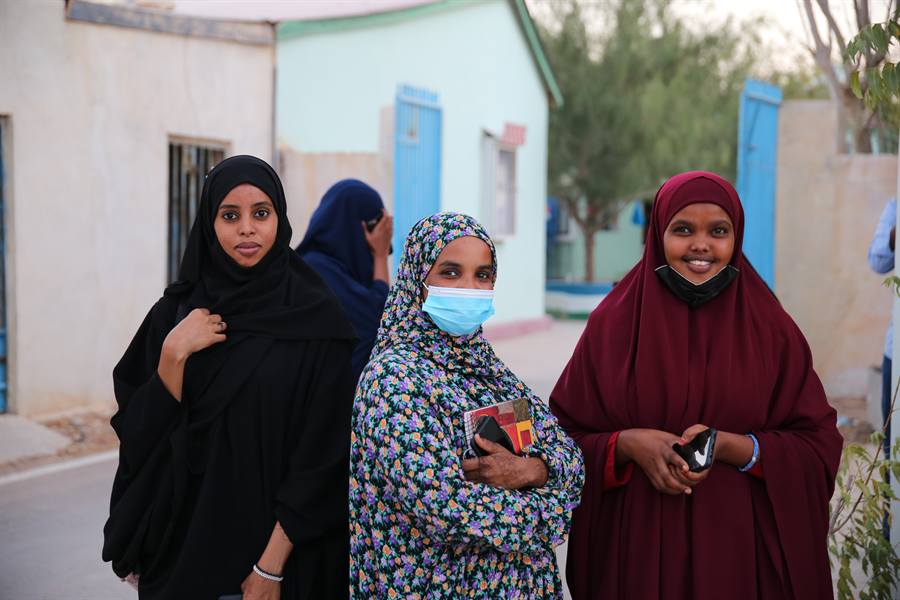 Three young women students carrying notebooks and phones smile outside their training institute.